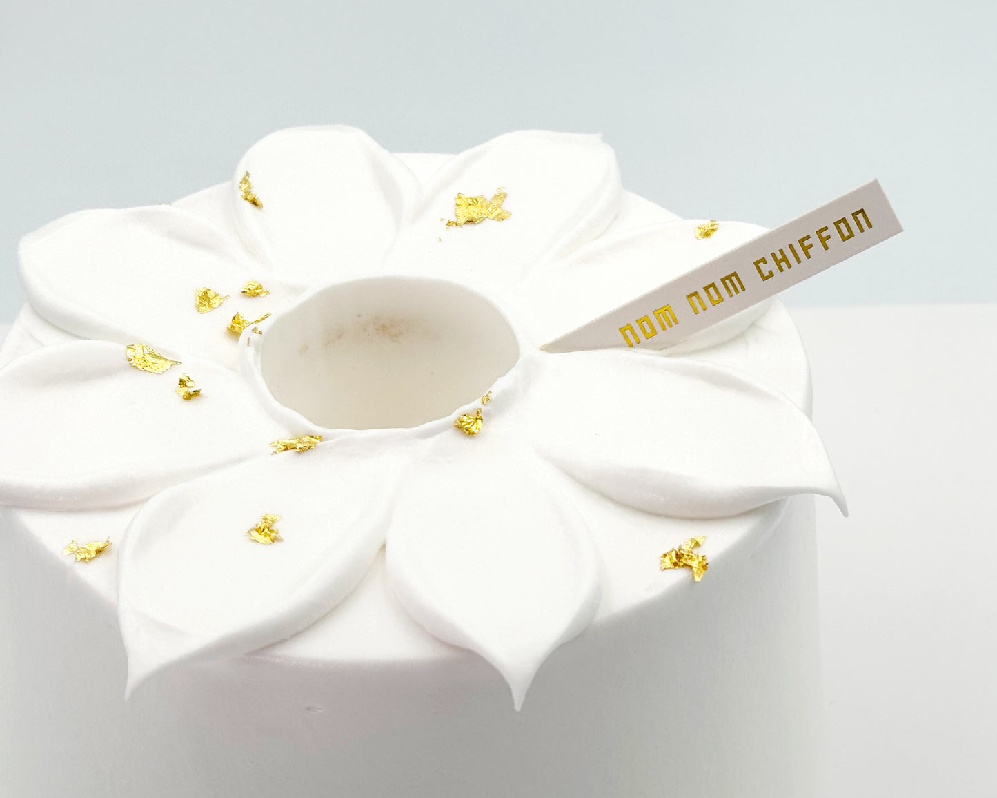 White frosted chiffon cake with flower petals and gold decorations and 'nom nom chiffon' label on a light gray background