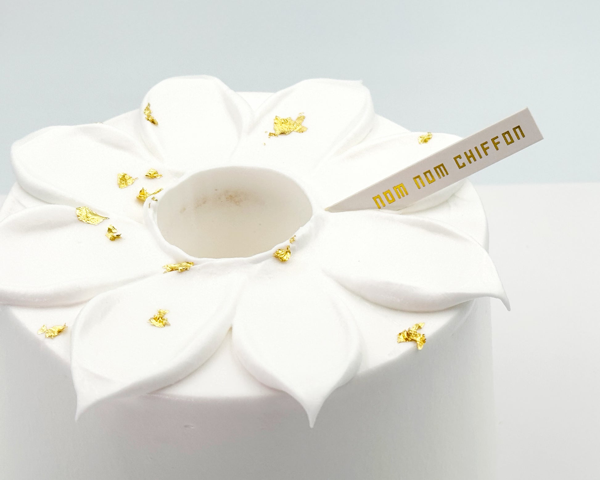 White frosted chiffon cake with flower petals and gold decorations and 'nom nom chiffon' label on a light gray background