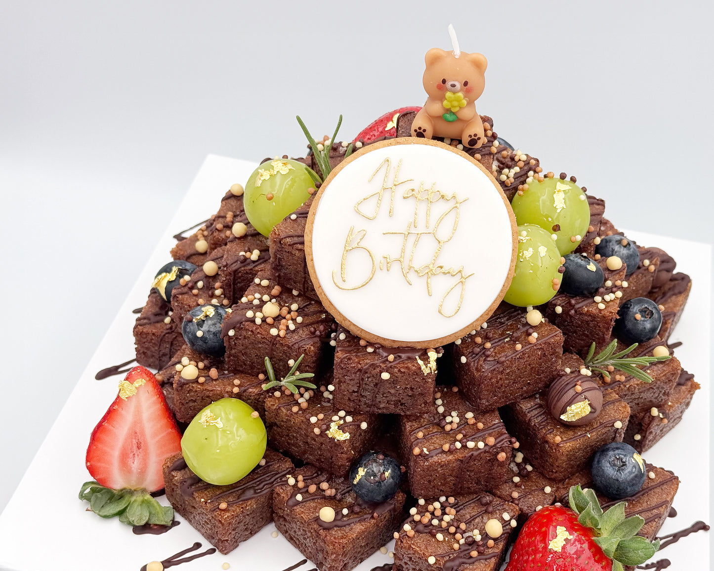 Fruit and brownie stacked cake with a 'Happy Birthday' candle on a white background