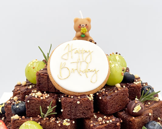 Brownie stacked cake with 'Happy Birthday' cookie and teddy bear candle on a light gray background