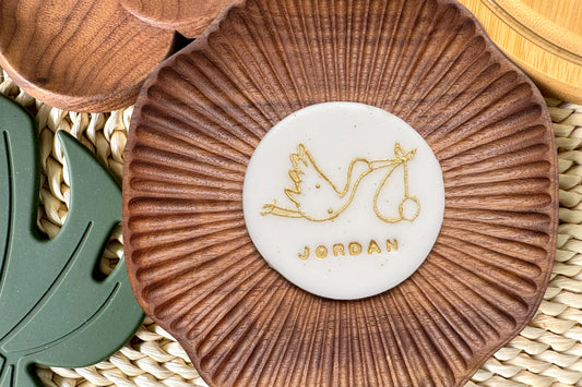 Brown textured coaster with a white cake topper featuring a stork carrying a baby and 'JORDAN' text, placed on a woven surface.