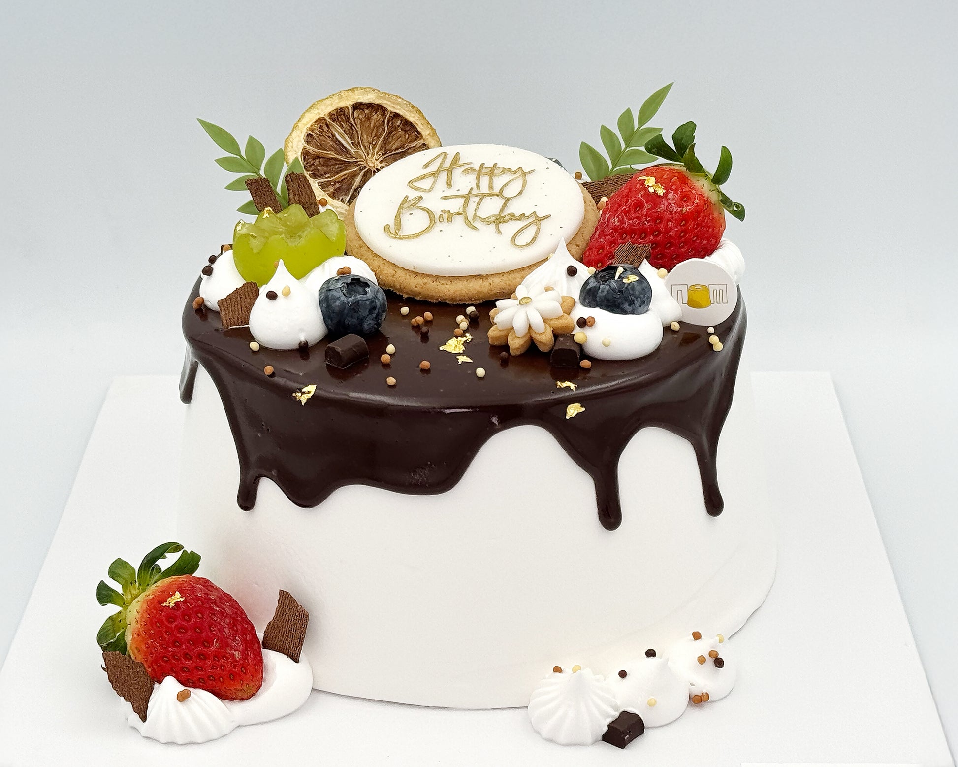 Decorative birthday cake with chocolate ganache, fruits, and a 'Happy Birthday' topper on a white background.