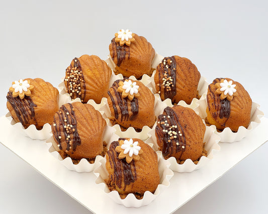 Decorative madeleines with chocolate drizzle and white flowers cookies on a white tray.
