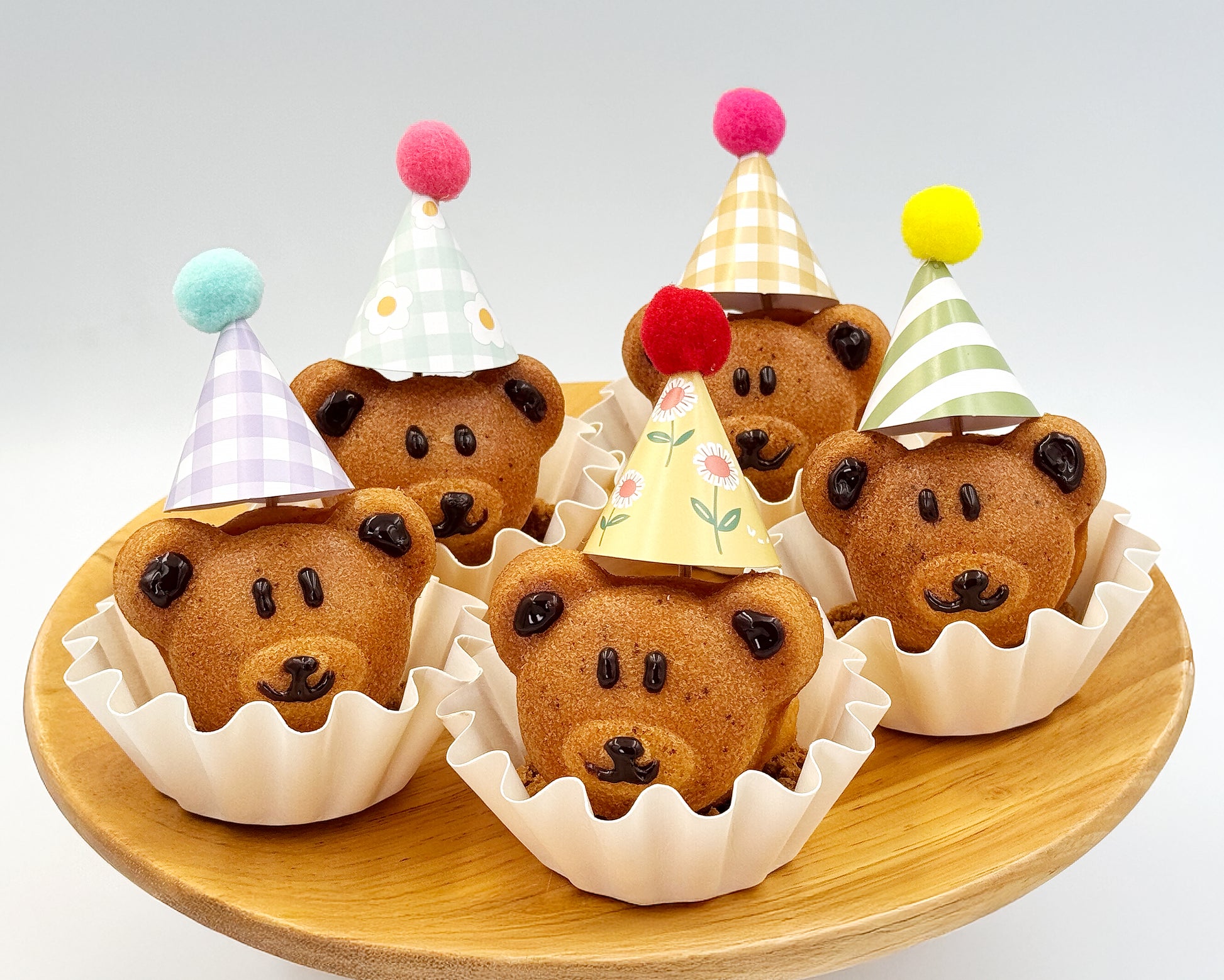 Bear-shaped madeleines with colorful party hats on a wooden plate.