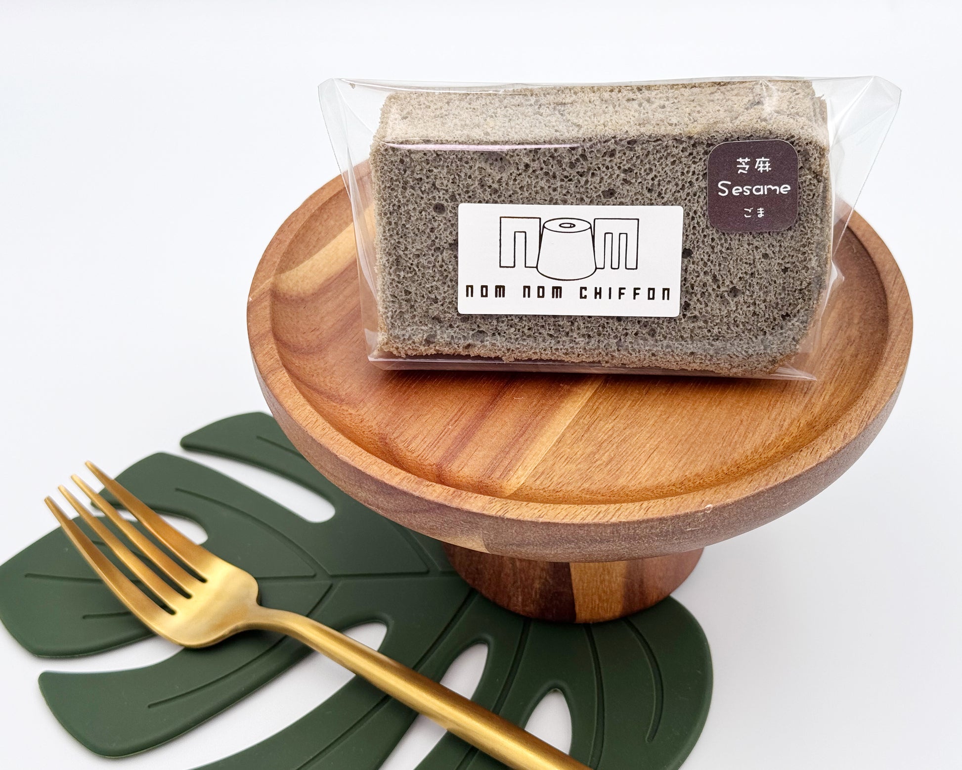 Packaged black sesame chiffon cake slice on a wooden stand with a gold fork and green leaves in the background