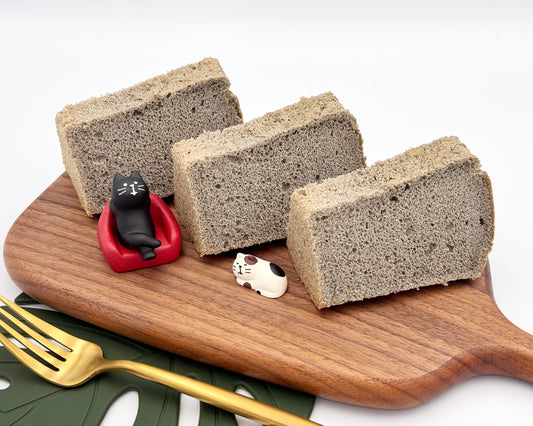 Three slices of black sesame chiffon cake on a wooden board with small cat figurines.