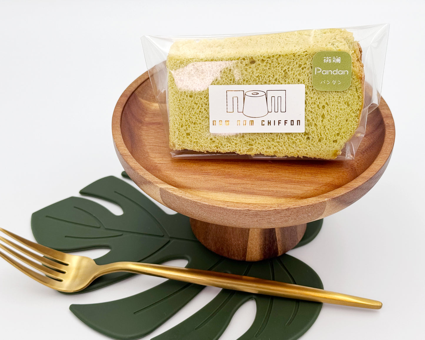 Packaged pandan chiffon cake on a wooden stand with a gold fork and green leaves on a white background