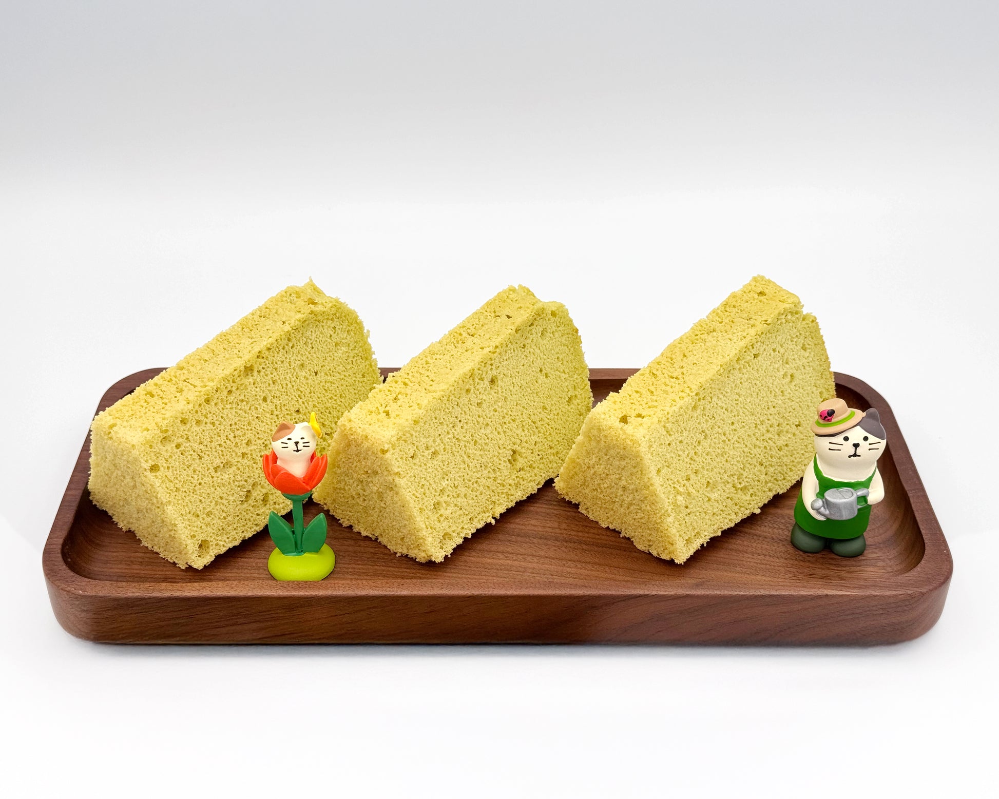 Three slices of pandan chiffon cakes on a wooden tray with small figurines on a white background