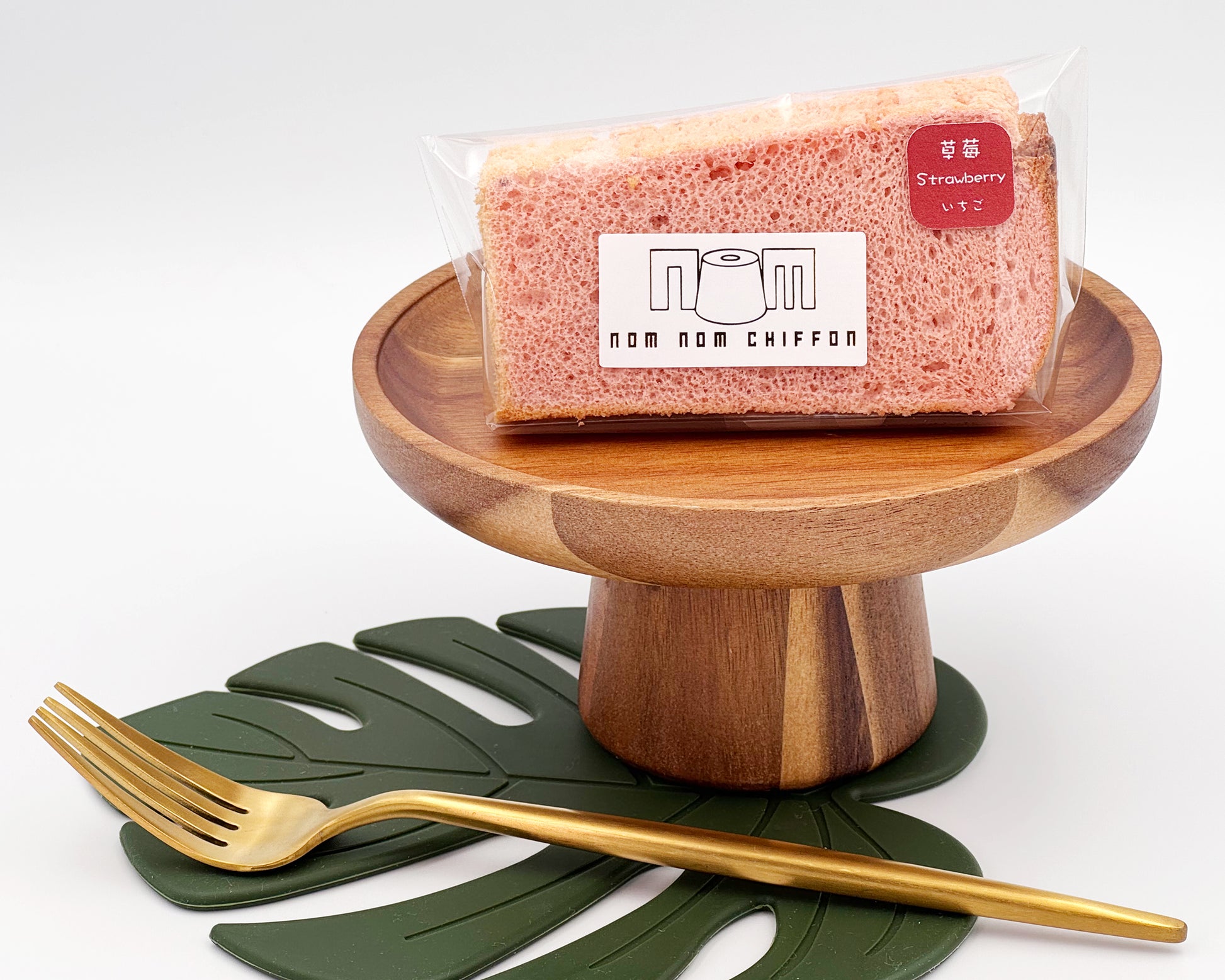 Packaged strawberry chiffon cake slice on a wooden stand with a gold fork and green leaf placemat.