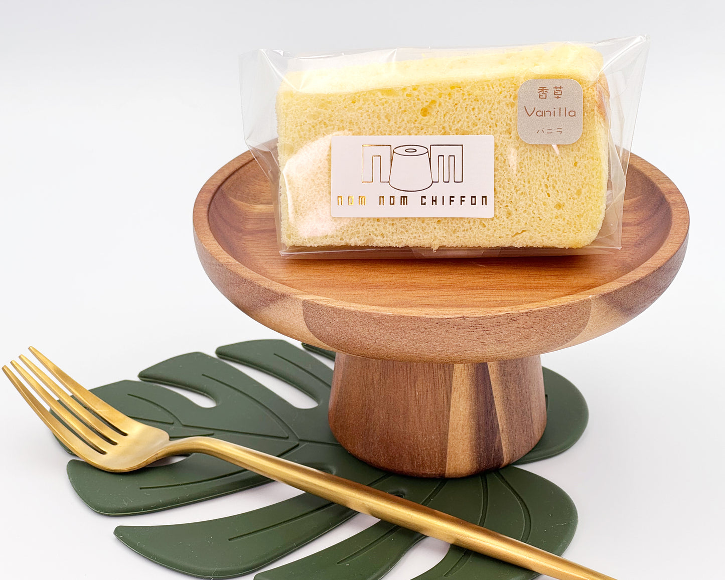 Packaged vanilla chiffon cake slice on a wooden stand with a gold fork and green leaf placemat.