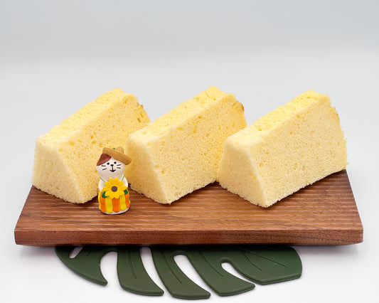 Three slices of vanilla chiffon cake on a wooden board with a small figurine and green leaf-shaped coaster.