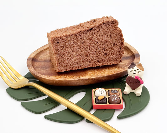 Slice of valrhona chocolate cake on a wooden plate with a gold fork, small decorative box, and figurine on a white background.
