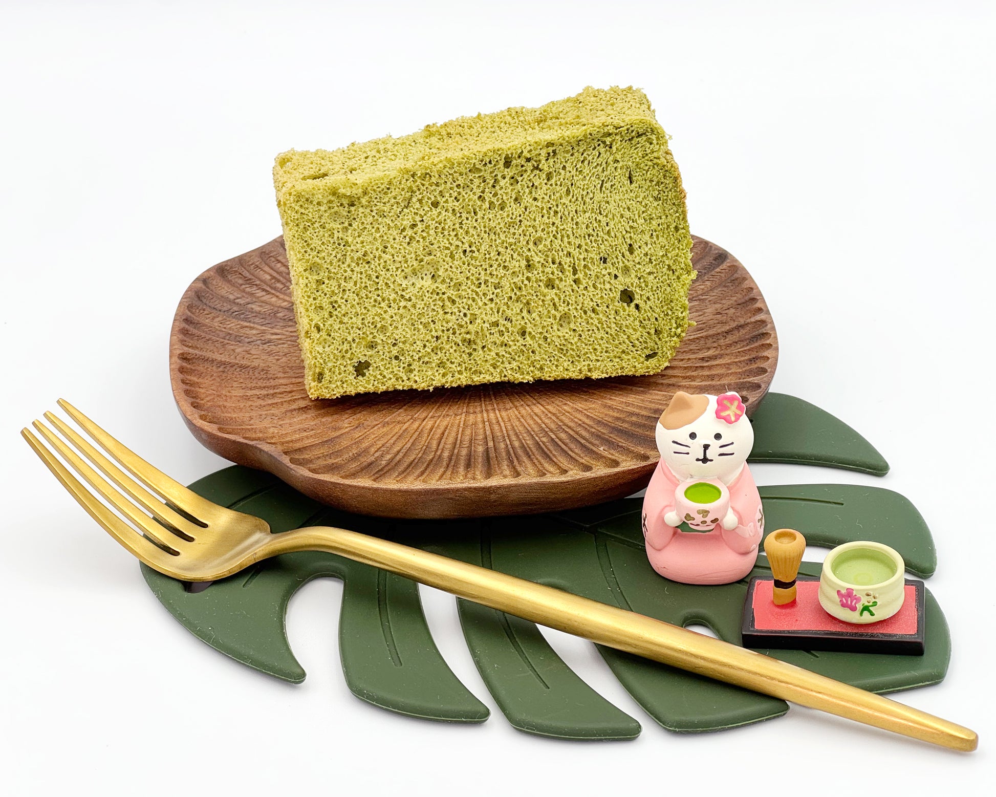 Premium matcha chiffon cake slice on a wooden plate with a cat figurine and tea set on a leaf-shaped mat.
