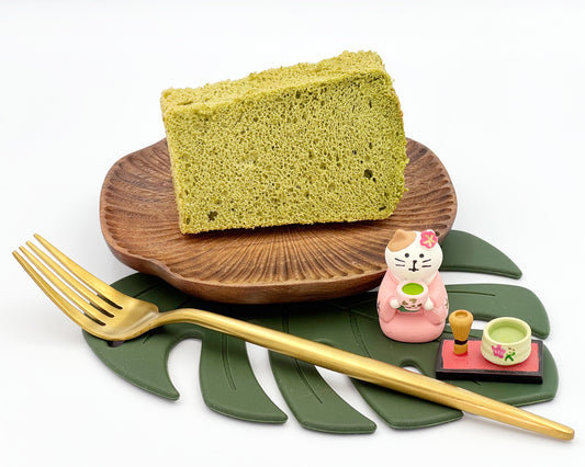 Premium matcha chiffon cake slice on a wooden plate with a cat figurine and tea set on a leaf-shaped mat.