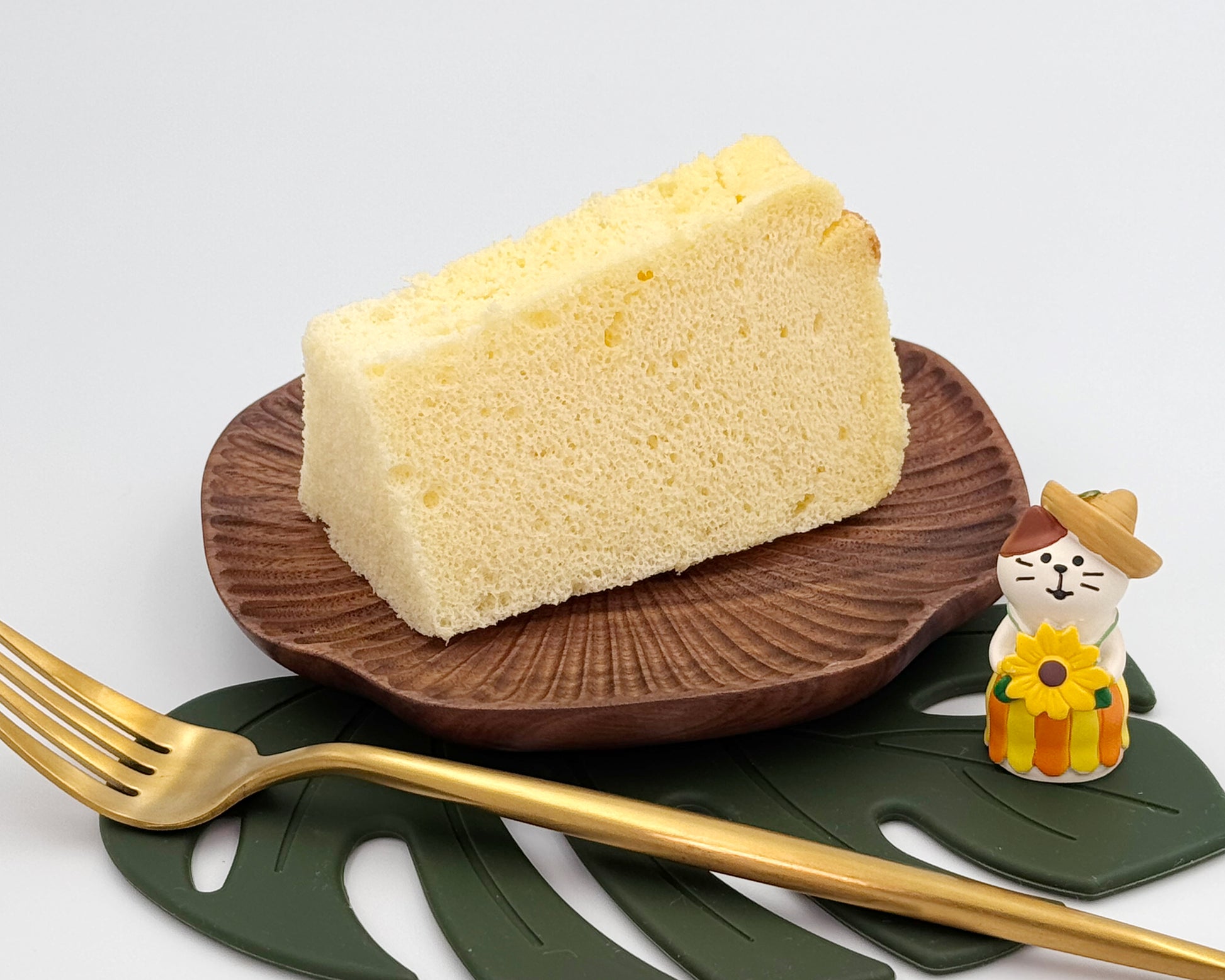 Slice of vanilla cake on a wooden plate with a gold fork and decorative figurine on a white background