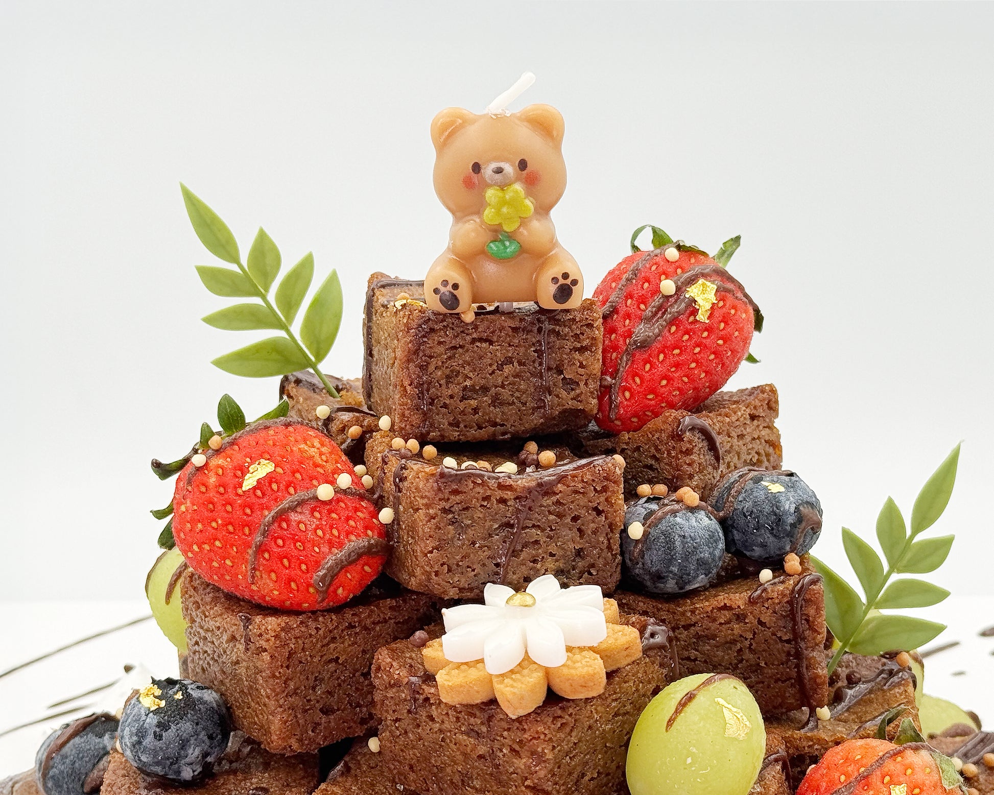 Stack of brownies with decorative elements including strawberries, blueberries, and a small bear figurine on a white background.