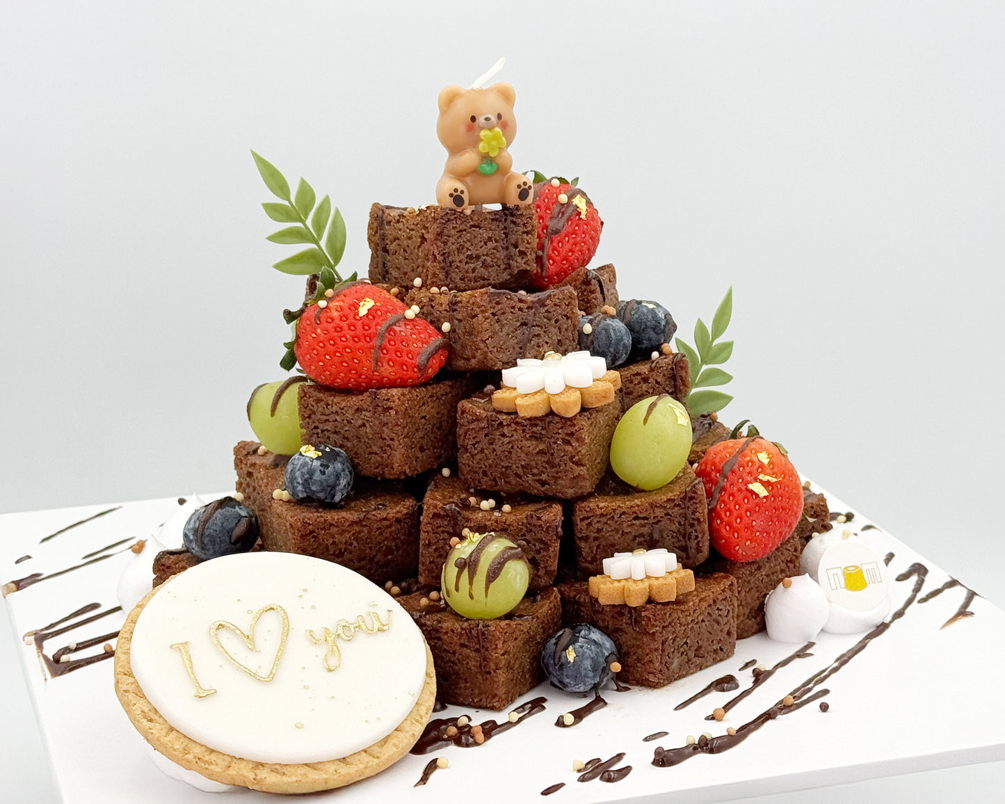 Decorative brownie stacked cake with fruits, and a small bear figurine on a white background.