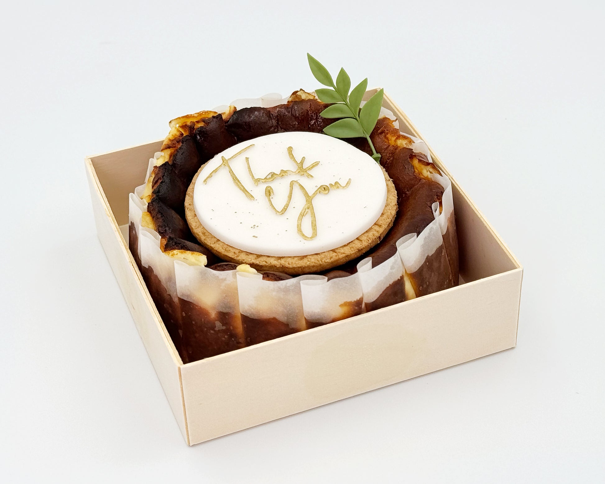 Basque burnt cheesecake in a box with 'Thank you' cake topper on it, on a light background
