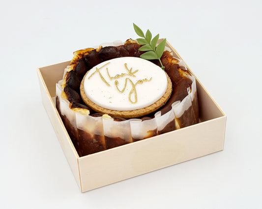 Basque burnt cheesecake in a box with 'Thank you' cake topper on it, on a light background