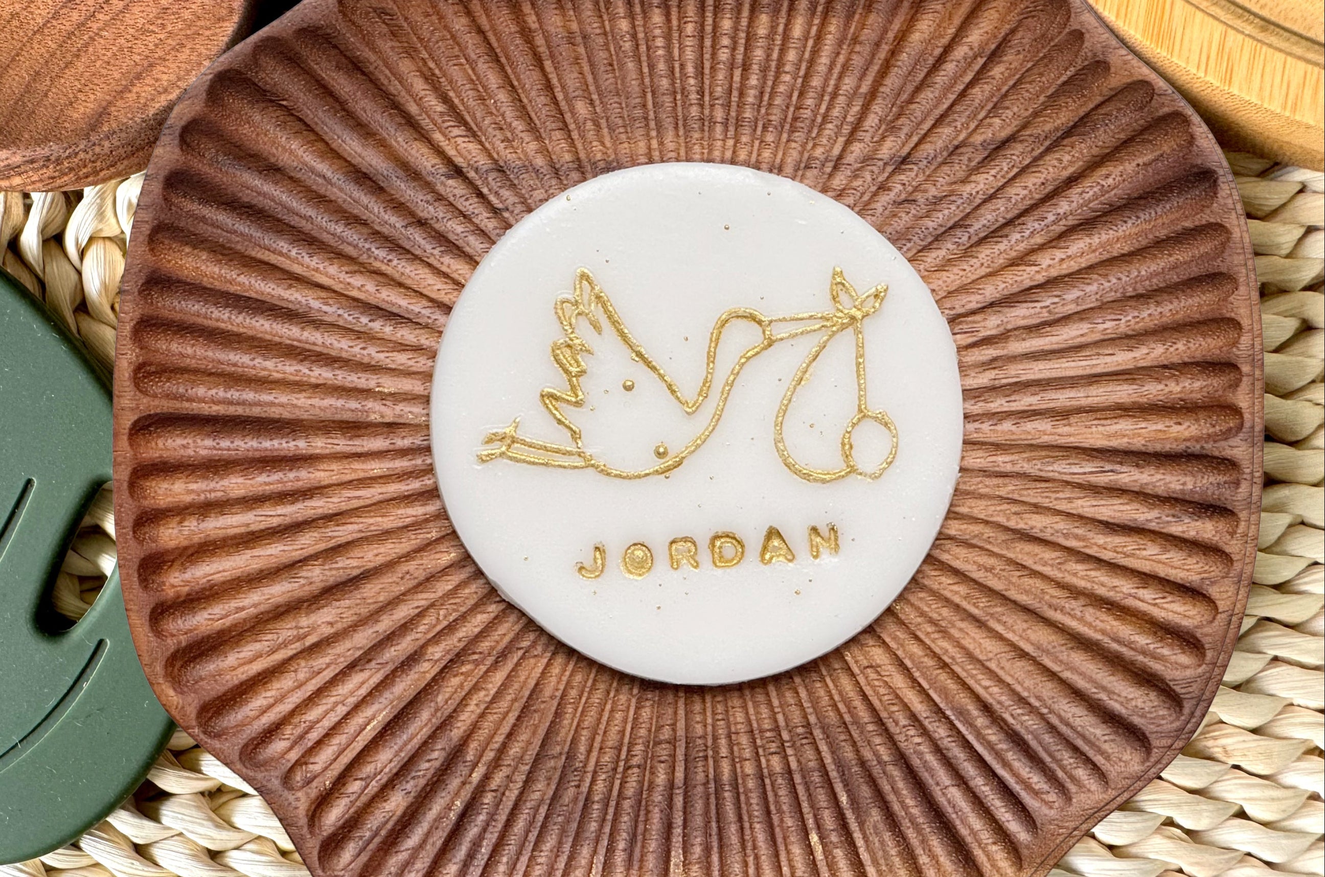 Stork carrying a baby and 'JORDAN' text on a cake topper, placed on a woven surface.