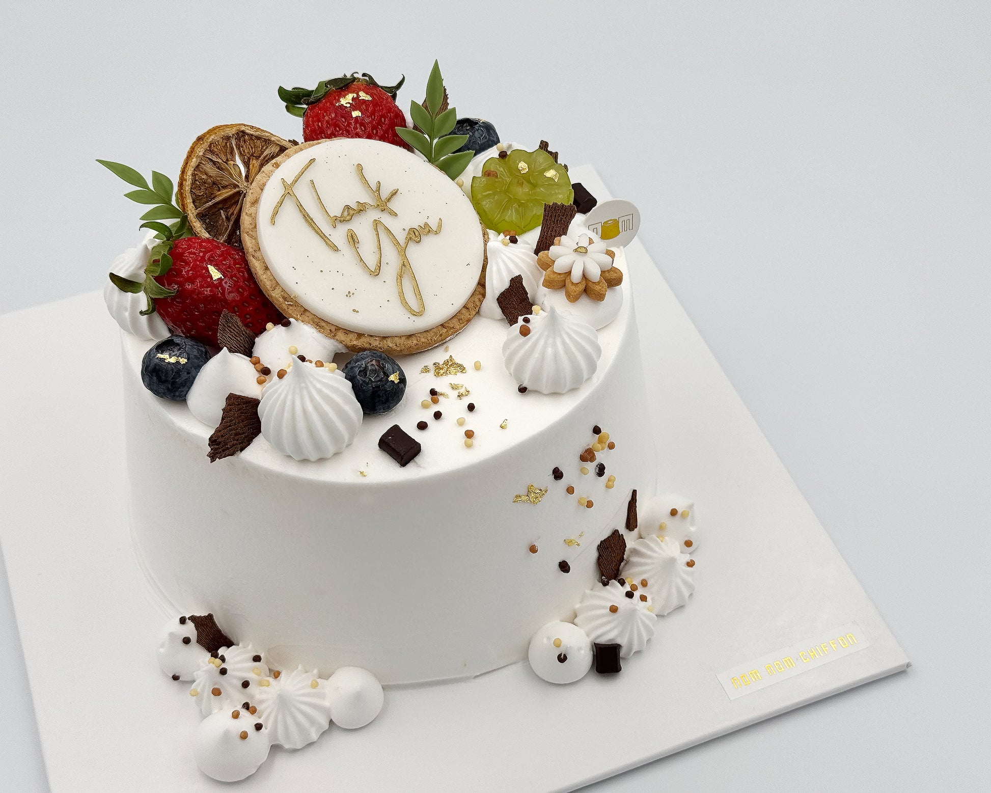 Side view of decorative chiffon cake with fruits and a cookie with 'Thank You' on a white background