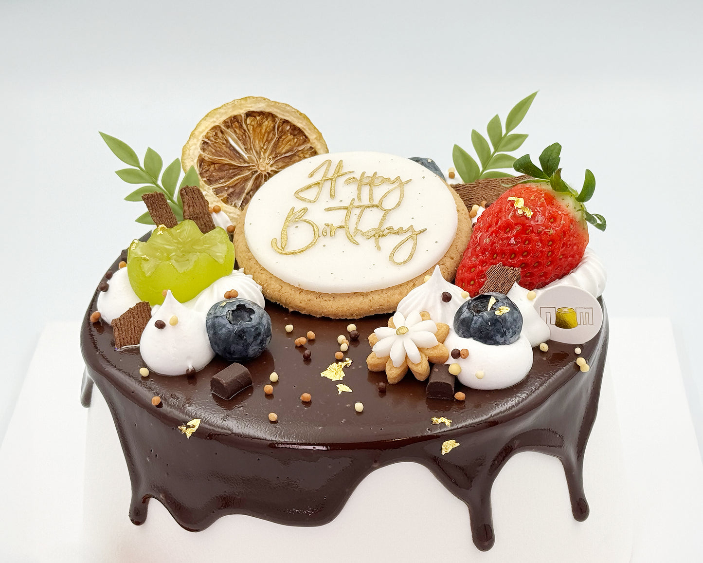 100% frosted chiffon birthday cake with chocolate frosting, fruits, and a 'Happy Birthday' cookie on a white background