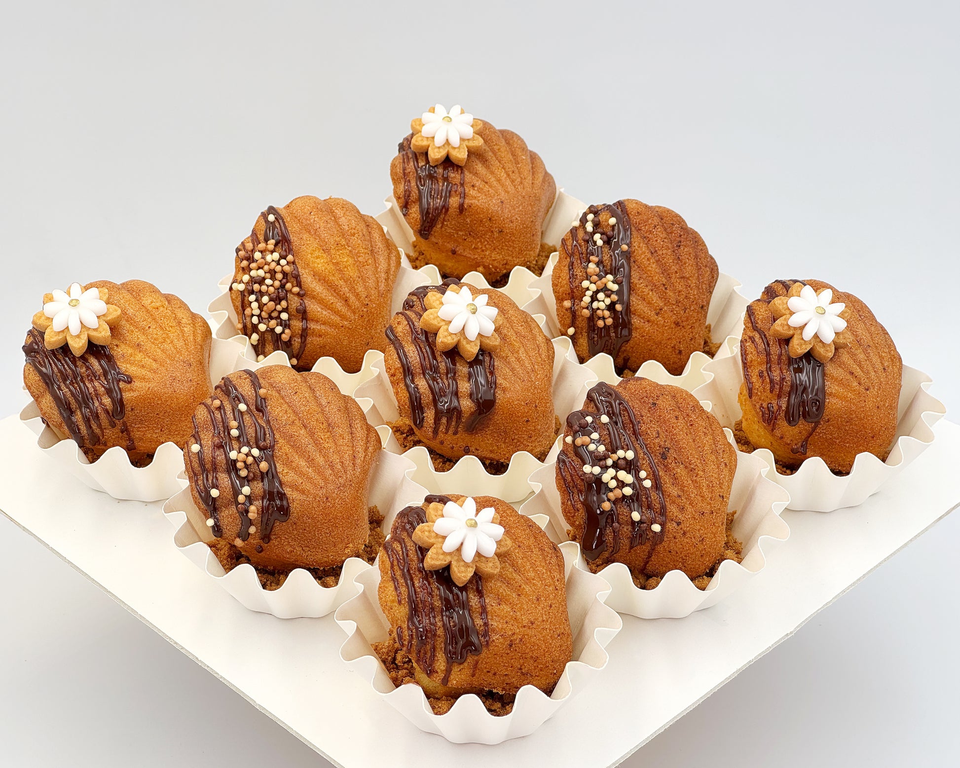 Decorative madeleines with chocolate drizzle and white flowers cookies on a white tray.
