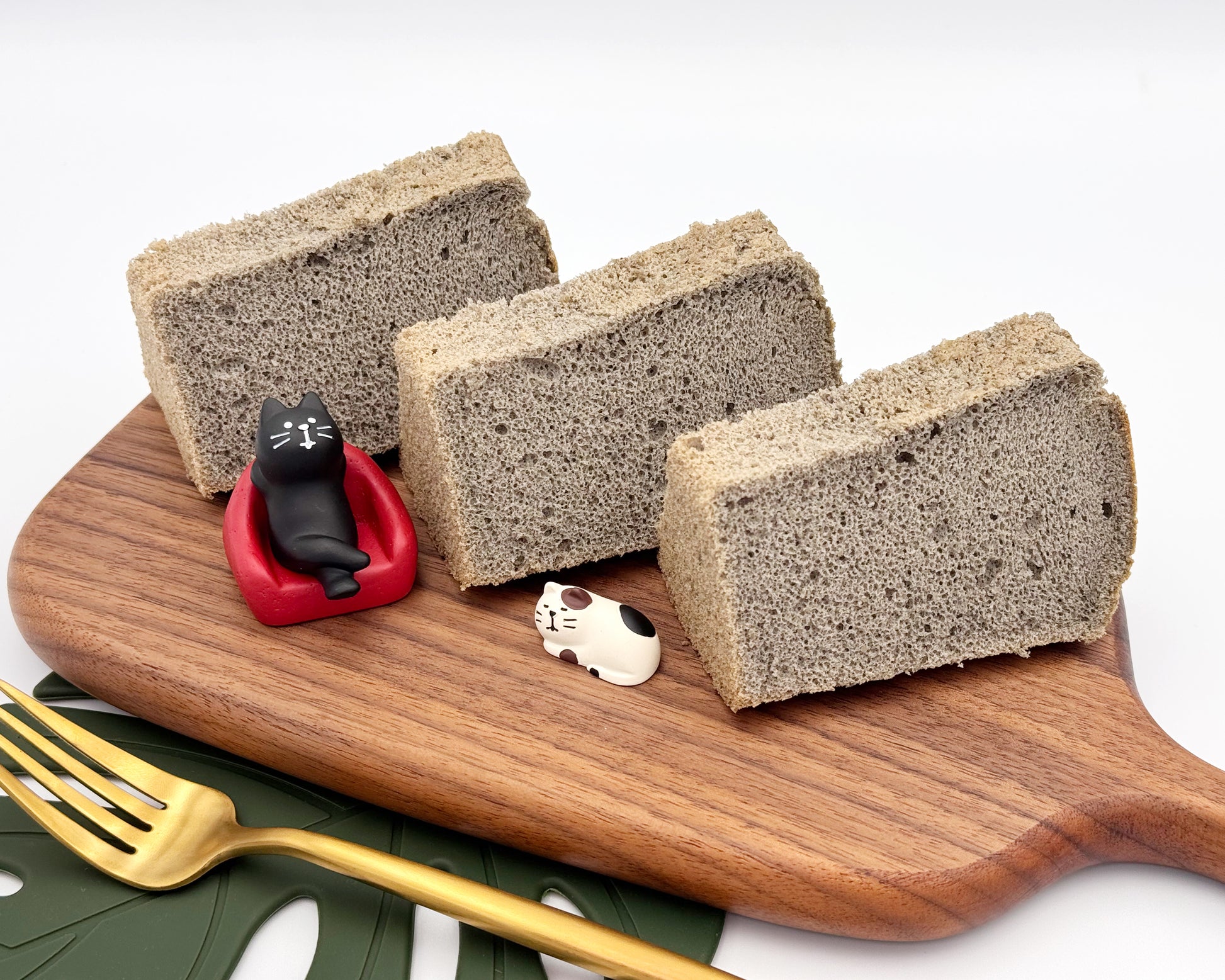 Three slices of black sesame chiffon cake on a wooden board with small cat figurines.