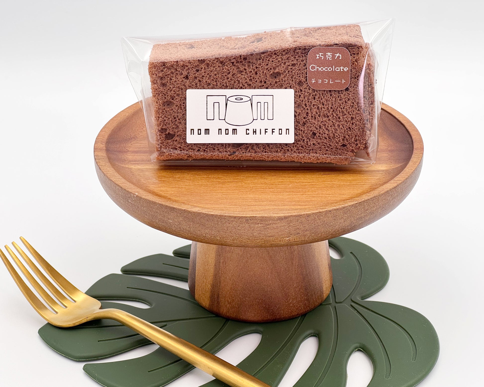 A slice of chocolate chiffon cake in packaging on a wooden stand with a gold fork and green leaf mat.