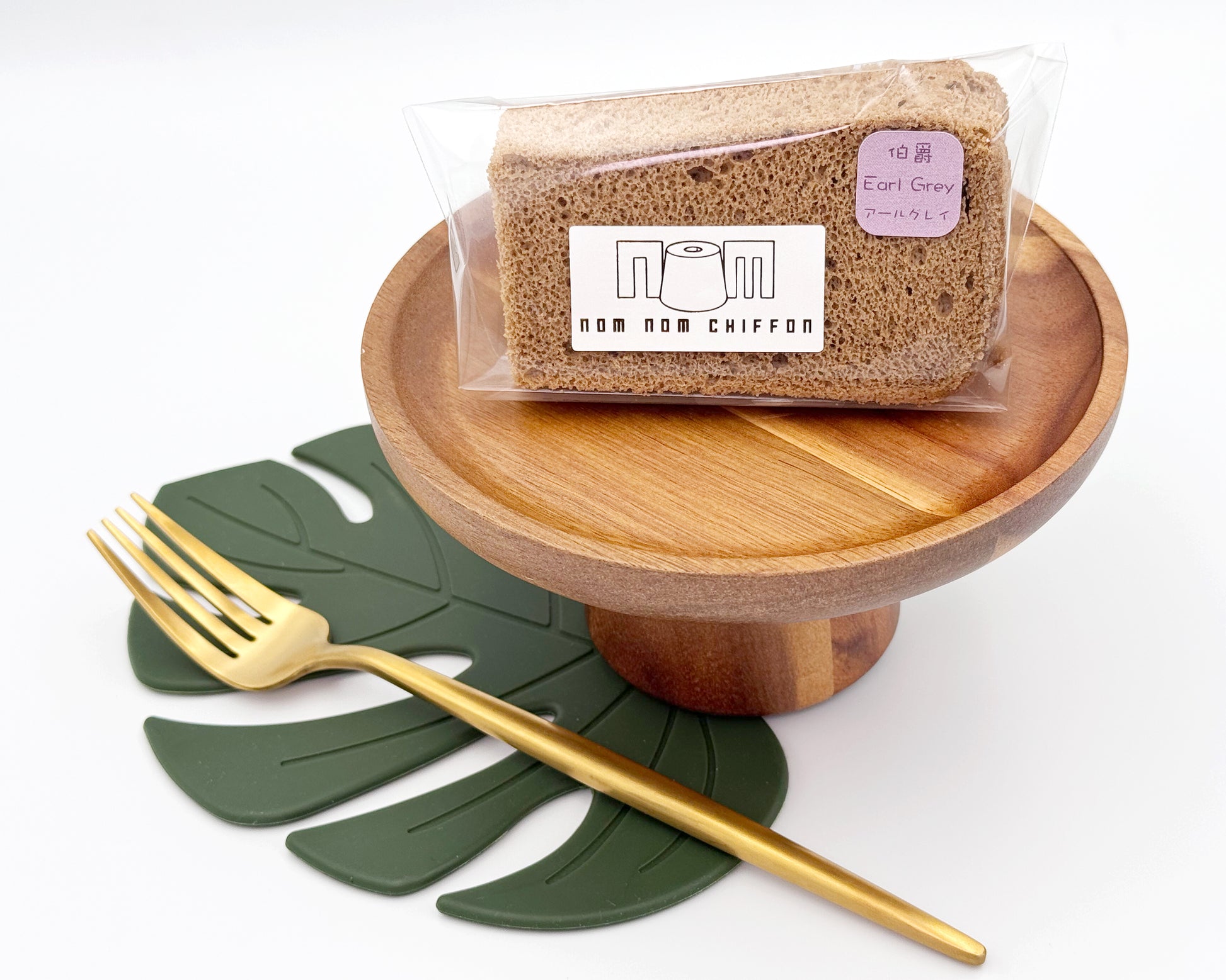 Packaged earl grey chiffon cake slices on a wooden stand with a gold fork and green leaf placemat.