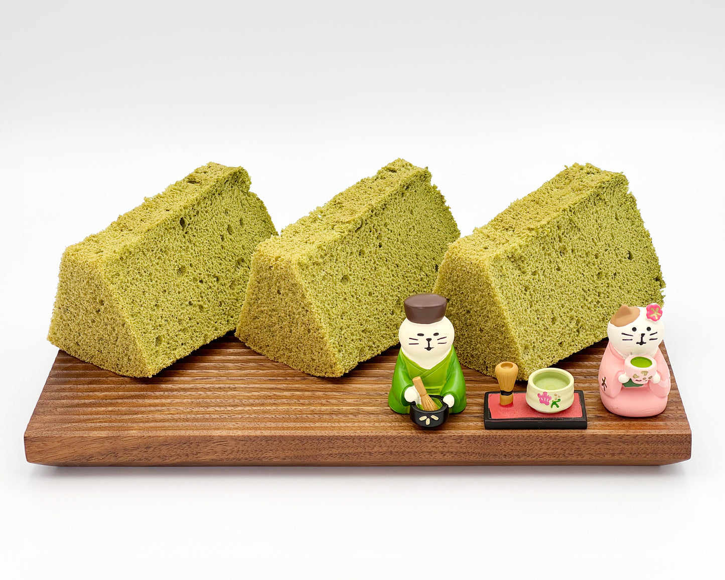 Three slices of premium matcha chiffon cake on a wooden board with small decorative figures on a white background