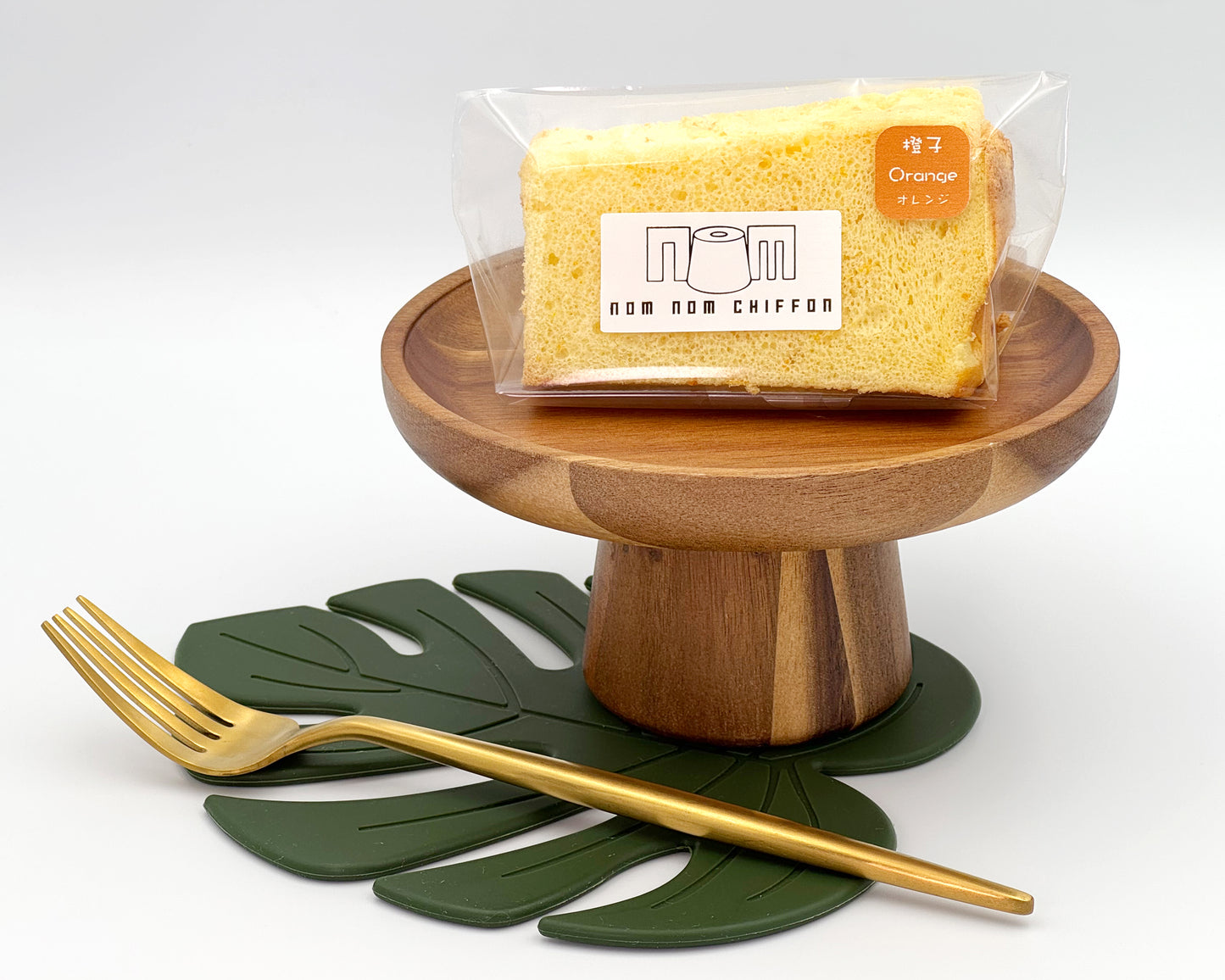 Packaged orange zest chiffon cake slice on a wooden stand with a gold fork and green leaf mat on a white background