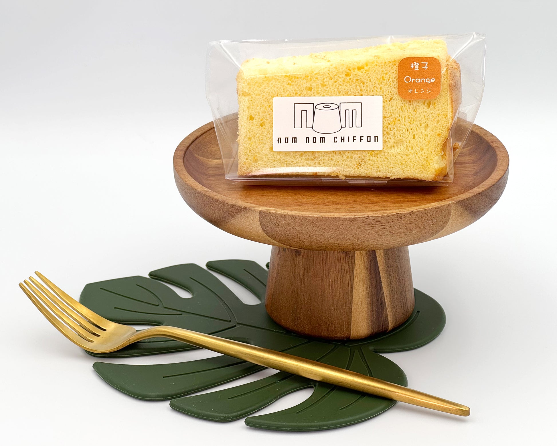 Packaged orange zest chiffon cake slice on a wooden stand with a gold fork and green leaf mat on a white background