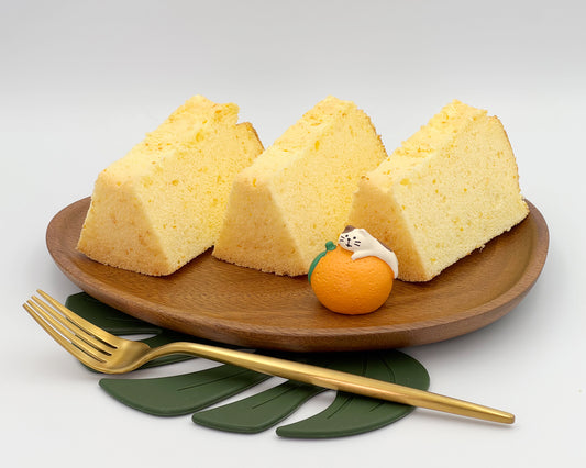 Three slices of orange zest chiffon cake on a wooden plate with a small decorative item and a gold fork.