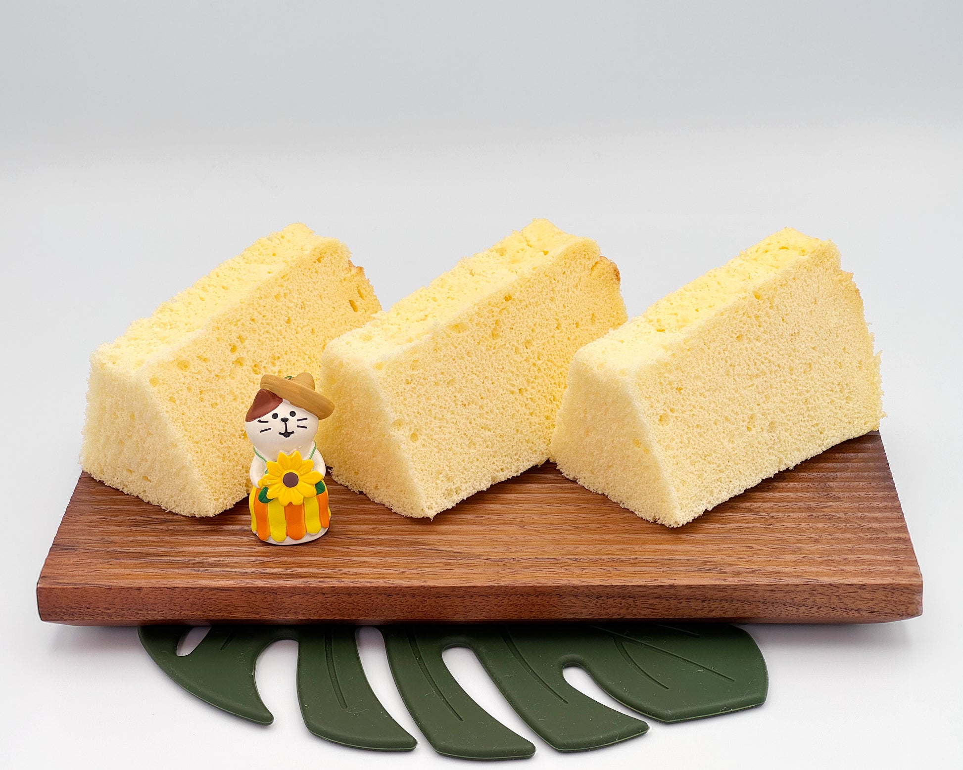 Three slices of vanilla chiffon cake on a wooden board with a small figurine and green leaf-shaped coaster.