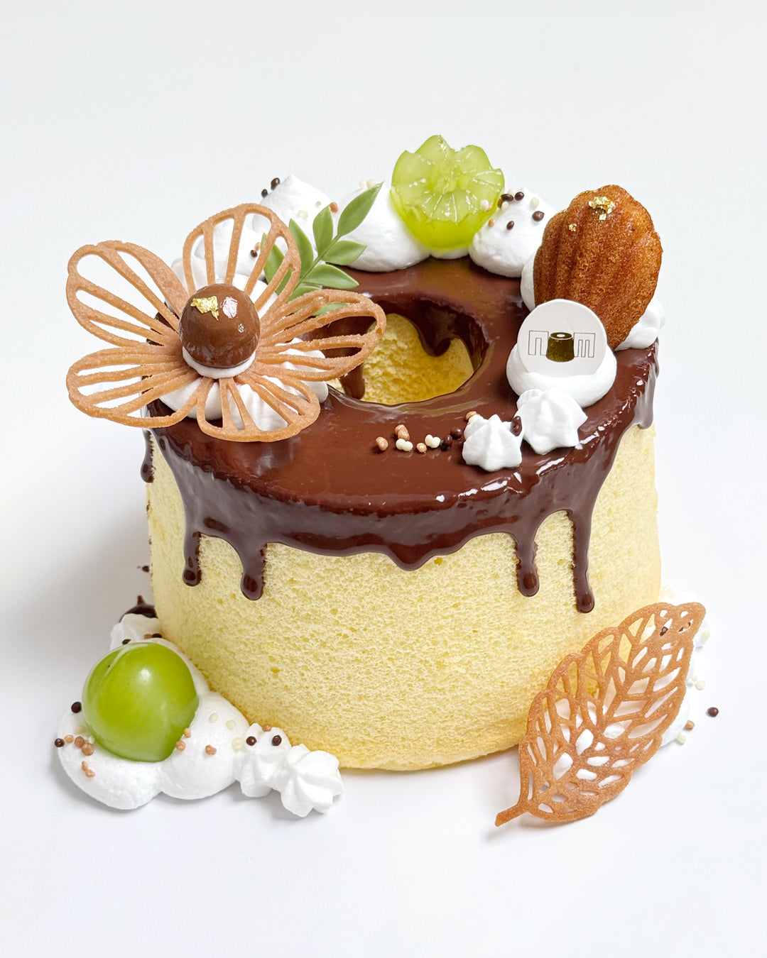 Decorative cake with chocolate ganache, flower tuile, mini madeleines, cookies, and fruits on a white background