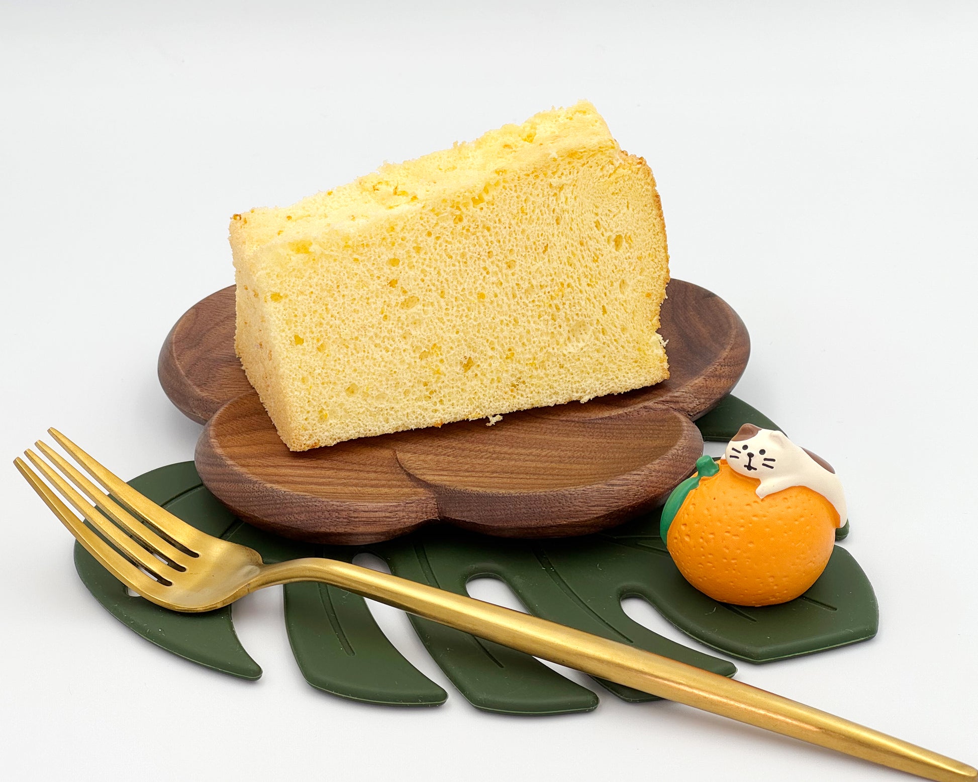 Slice of orange zest chiffon cake on a wooden plate with a gold fork and decorative elements on a white background